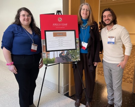 Three people stand indoors beside a sign on an easel for the Adele H. Stamp Student Union at the University of Maryland. From left to right: Nicole Holt, a public health major and disability studies minor; Andrea Kalfoglou, PhD, Associate Professor of Public Health; and Michael Berardi, Assistant Director of Retriever Essentials. All are smiling and wearing conference name badges.