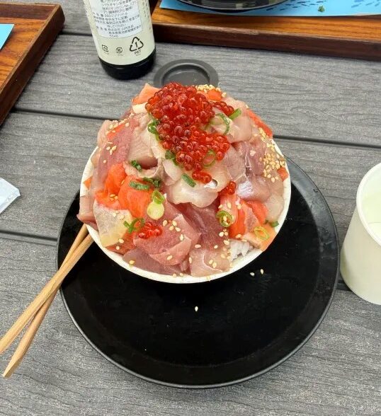 A high-angle shot of a Japanese kaisen-don (sushi rice bowl) overflowing with a large mound of assorted diced raw fish, topped with bright red ikura (salmon roe), sesame seeds, and green onions.