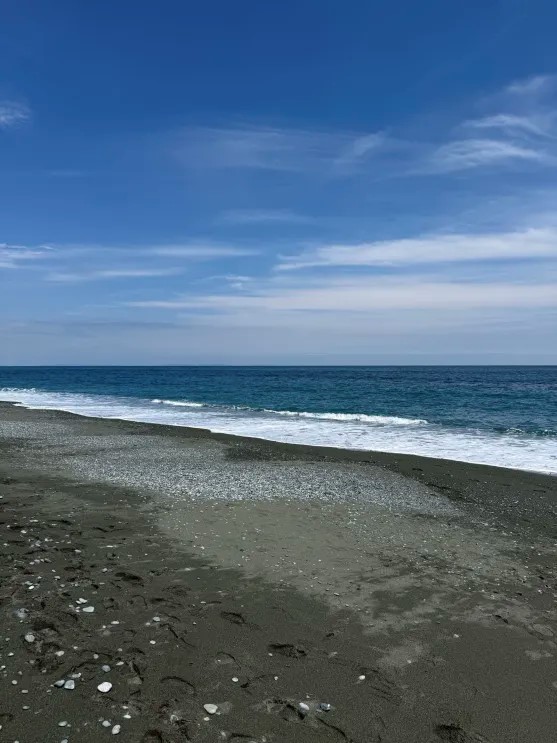A portrait-oriented wide shot of an empty, dark sand beach meeting a deep blue ocean under a bright sky with light, wispy clouds.