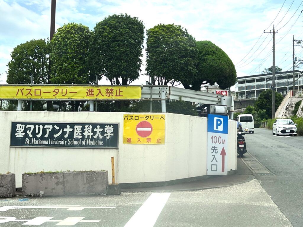 A photo showing the entrance to St. Marianna University School of Medicine. Detailed signage in Japanese and English is clearly legible on a retaining wall, with manicured trees above and a street scene to the right.