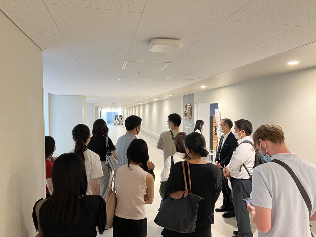A group of masked visitors stands in a bright, modern hospital corridor outside a doorway marked '6D Rehabilitation Center,' listening to two guides in business attire.