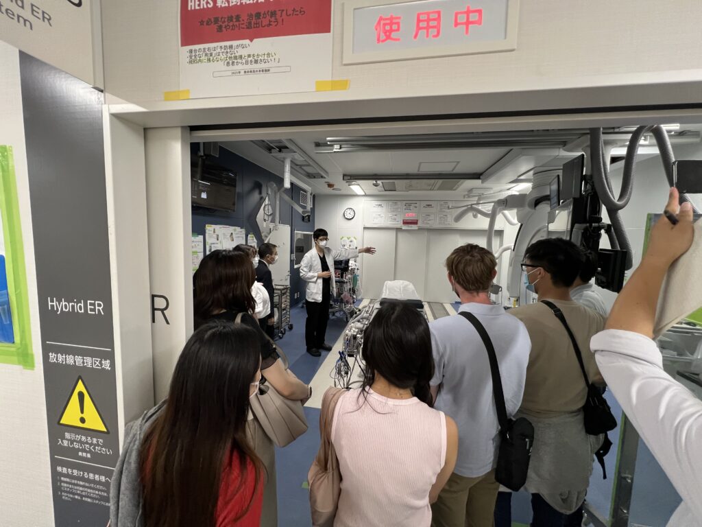 A group of masked visitors stands at the entrance of a Hybrid ER room, listening to a medical professional in a white lab coat who is gesturing toward advanced medical equipment inside.