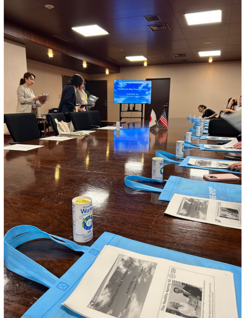 A perspective photograph taken from one end of a very long, highly polished wooden conference table in a formal meeting room at the Kanagawa Prefectural Government building (Untitled design (1).jpg). The image captures the setup for the meeting between study abroad students and Governor KUROIWA Yuji described in the blog. Numerous individual settings for attendees line the table, each featuring a blue reusable tote bag printed with the concept "ME-BYO" and topped with a printed presentation packet titled "Healthcare New Frontier Policy" with a photo of the Governor. Paper water cartons are also present. In the background, a large screen displays the presentation slide: "Healthcare New Frontier Policy" and "KUROIWA Yuji, Governor of Kanagawa Prefecture". Small Japanese and US flags are placed centrally on the table. Several staff members are standing and working on the left, and students are seated on the right. This scene physicalizes the discussion about public policy and the 'ME-BYO' concept mentioned in the blog text.