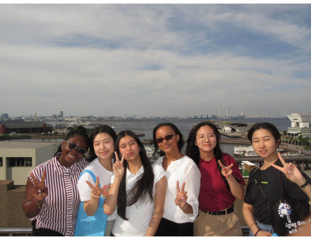 A group of six study abroad students pose together on an observation deck overlooking Yokohama Harbor in Kanagawa Prefecture, Japan. Smiling and making peace signs, the students stand with the waterfront, bridges, and city skyline in the background following a day of learning about aging policy and cultural exchange.