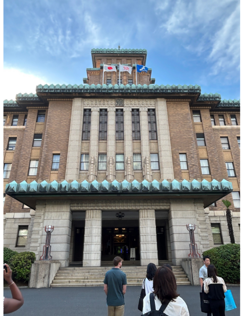 Front view of the Kanagawa Prefectural Government building in Japan, an imposing historic brick-and-stone structure with Art Deco architectural details and a green copper roof. Japanese and prefectural flags fly above the entrance under a blue sky with wispy clouds, while several visitors stand in front of the building near the main staircase.