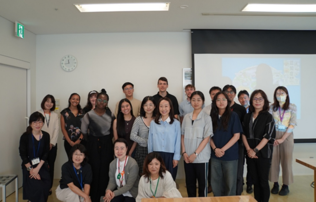 A group photo of approximately 20 individuals of diverse ethnicities and genders posing in a bright indoor room, likely a classroom or meeting space. Three women are kneeling in the front row, while the remaining participants stand in rows behind them, dressed in casual and smart-casual clothing. The background features a white wall with an analog clock, an exit sign on the far left, and a large projection screen displaying a colorful graphic on the right.
