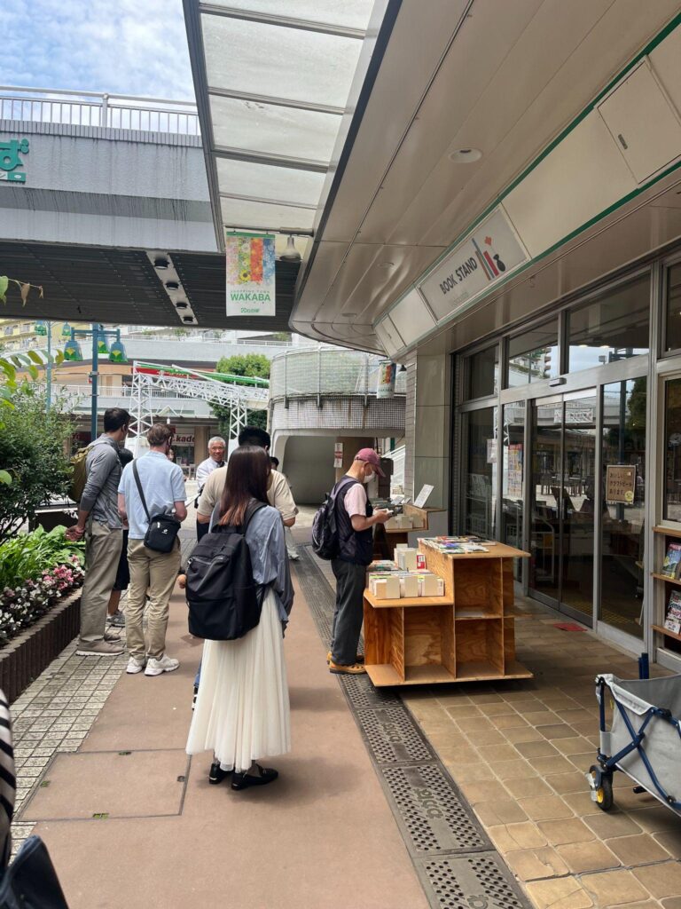 A group of people standing outside the 'BOOK STAND Wakabadai' shop, browsing an outdoor wooden book display under a covered walkway.