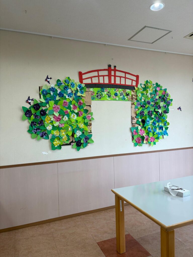 Colorful wall display inside the Yoko-No-Sono nursing home featuring handmade paper flowers arranged around a decorative red bridge. The artwork, created in bright shades of green, blue, pink, yellow, and purple, adds a cheerful and welcoming atmosphere to a shared resident space.