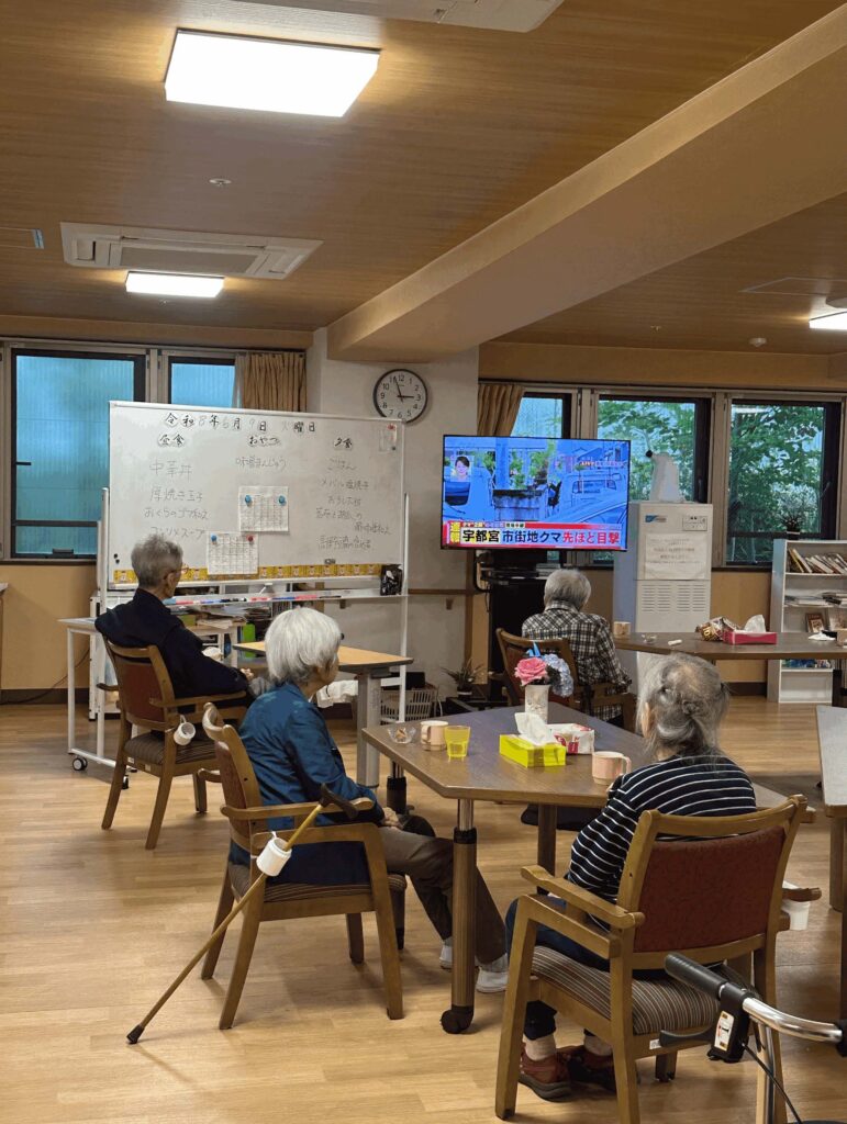 Elderly residents sit in a warmly lit common room watching a television news broadcast next to a whiteboard detailing the daily menu at a senior care facility.