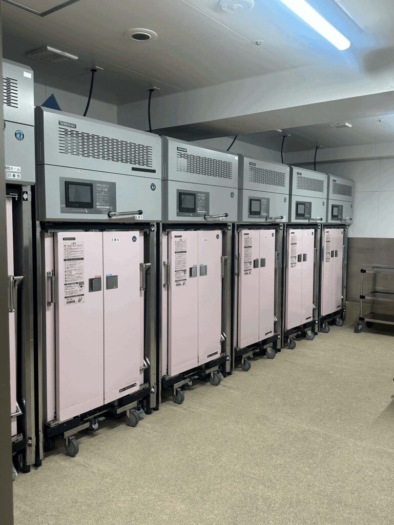A row of industrial Hoshizaki food service or medical warming cabinets with light pink double doors on wheels, lined up under large silver ventilation units in a commercial facility.
