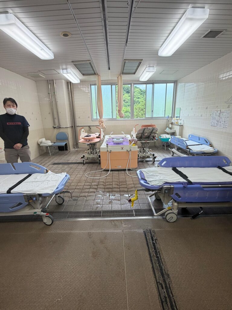 An institutional assisted bathing room in a Japanese nursing home. Multiple specialized blue and pink mobile shower gurneys with safety straps are positioned around a central mechanical bathing unit. A staff member wearing a face mask stands on the left, and a large window in the background looks out onto green trees.