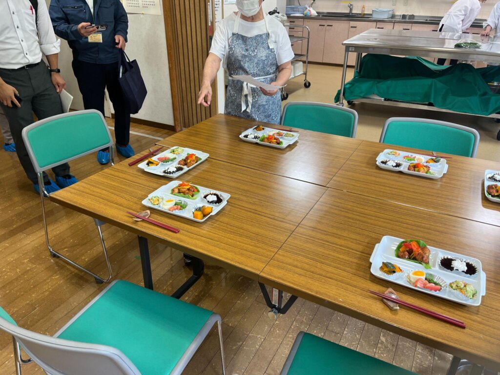 Prepared bento-style meal trays arranged on a wooden table in a community kitchen, while a masked instructor in an apron points toward one of the settings.