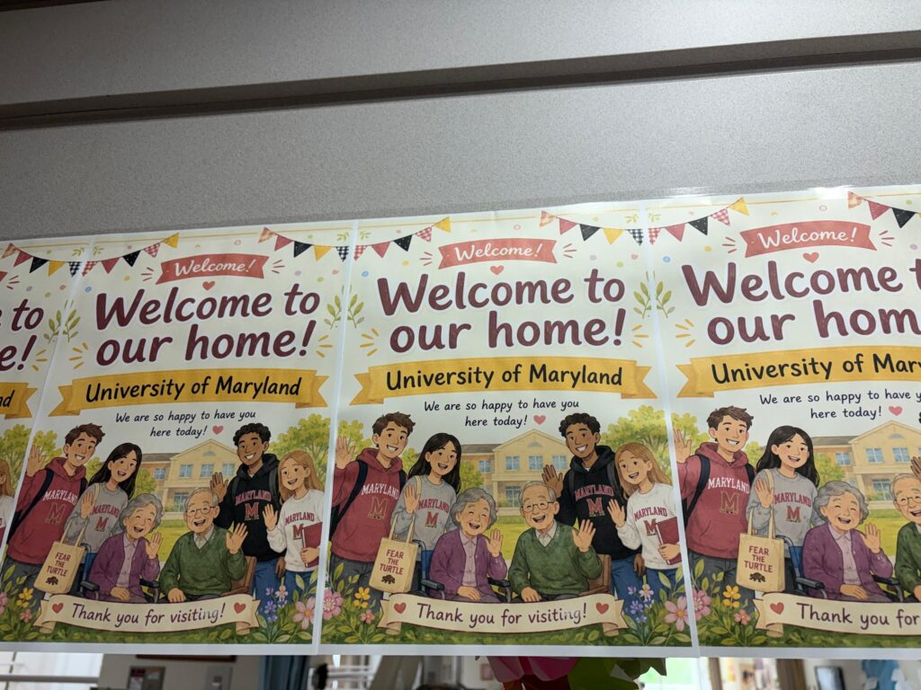A row of identical, repeating welcome posters hanging side-by-side. The posters feature the text "Welcome to our home! University of Maryland" above a colorful illustration of four smiling college students in Maryland apparel waving alongside an elderly couple, one of whom is using a wheelchair.