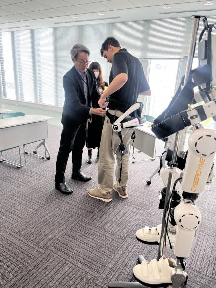 A staff member adjusts a lower-body robotic exoskeleton worn by a participant during a mobility demonstration, while a full humanoid assistive robot stands nearby in a bright rehabilitation training room.