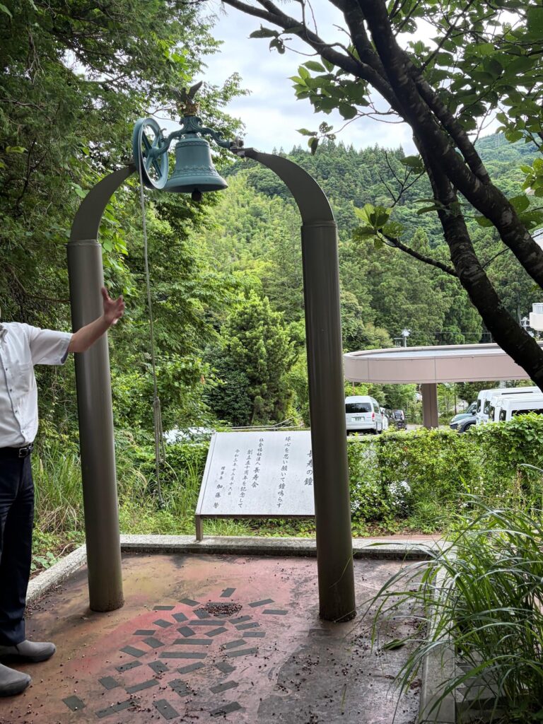 An outdoor commemorative bronze bell hanging from a large metal archway on a concrete platform. In the background, a stone monument plaque with Japanese inscriptions stands before a lush, forested hillside. On the left, a person's arm is raised, gesturing toward the bell.