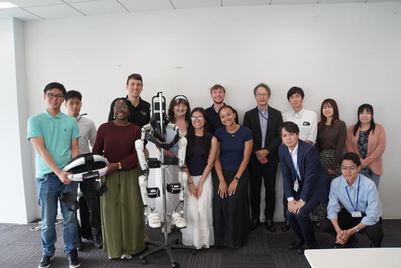 Group photo of students, faculty, and staff standing together with a wearable robotic exoskeleton on display during a visit to a rehabilitation robotics center.