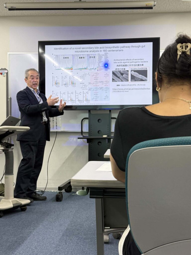 A presenter in a suit gestures toward a large digital screen displaying a scientific presentation titled "Identification of a novel secondary bile acid biosynthetic pathway through gut microbiome analysis in 160 centenarians." An audience member is visible in the foreground, viewed from behind.