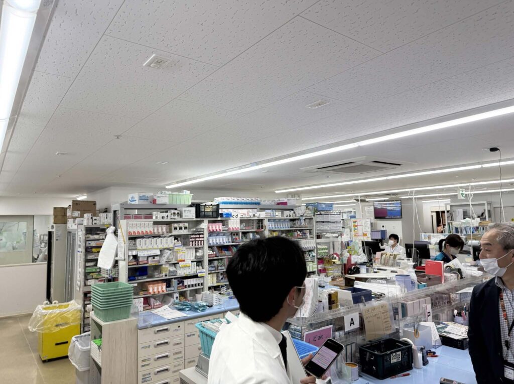 A busy, brightly lit pharmacy dispensary filled with rows of medication shelves. In the foreground, a staff member in a white lab coat holds a phone next to a man in a blazer; both are wearing face masks.