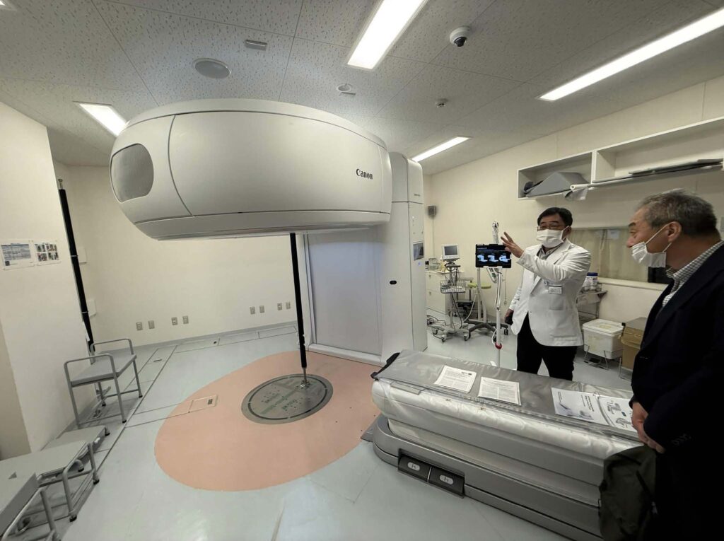 A medical professional in a white lab coat gestures toward a control screen next to a large Canon medical imaging machine while explaining it to a man in a dark blazer.